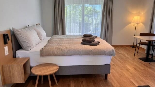 Zimmer im Elternhaus beim Kinderspital Zürich: Doppelbett mit beige gestreifter Bettwäsche, darauf gefaltete Frottiertücher in Taupe, dahinter ein Fenster mit Vorhängen