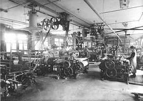 Historische Aufnahme eines Websaals mit mechanischen Webstühlen, angetrieben über Riemen von Wellen an der Decke