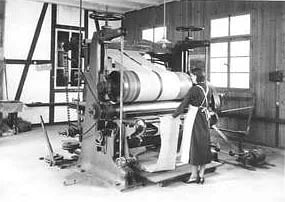 Historische Aufnahme: Arbeiterin an einer grossen Walzenmaschine, über die eine Stoffbahn läuft