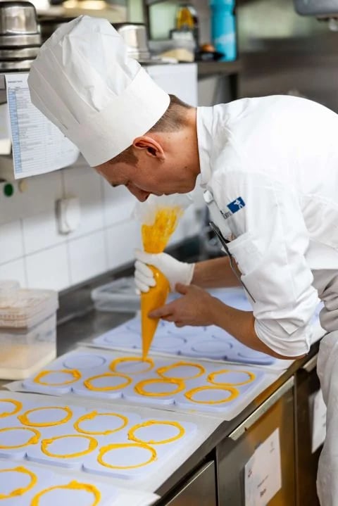 Koch mit Kochmütze spritzt mit dem Dressiersack orange Ringe auf eine Silikonmatte